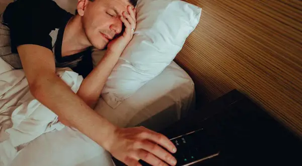 Man in bed, hand reaching for phone alarm. Does snoozing make you more tired?