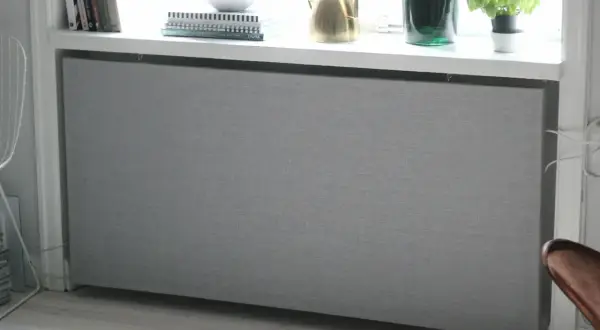 Grått elementkydd i tyg, ett DIY-projekt som ger ett stilrent intryck. Böcker och en vattenkanna står på fönsterbrädan ovanför.