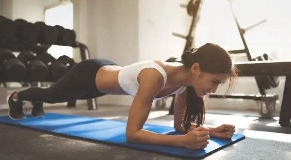 Woman performing a plank jack exercise on a blue mat in a gym, with weights in the background.