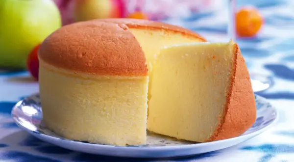 Una soffice torta di cotone, dorata e leggera, servita su un piatto con mele verdi e rosse sullo sfondo.