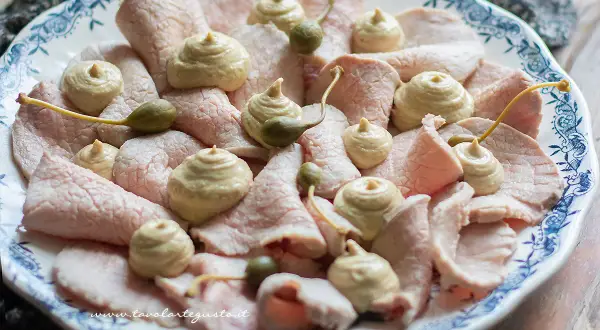 Vitello tonnato della nonna: fette sottili di carne rosa, guarnite con salsa tonnata e capperi, su un piatto bianco con decori blu.