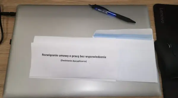 Na laptopie leżą dokumenty z napisem "Rozwiązanie umowy o pracę bez wypowiedzenia (Zwolnienie dyscyplinarne)". Obok długopis i myszka.