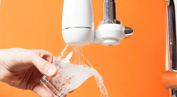 Un robinet avec un filtre blanc verse de l'eau dans un verre. C'est le moment de changer le filtre robinet pour une eau plus pure.