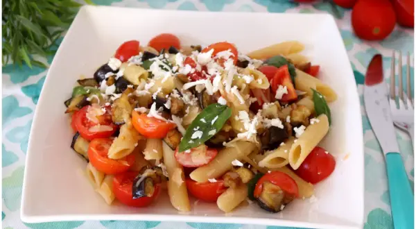 Un piatto di pasta fredda alla norma con melanzane fritte, pomodorini freschi e ricotta salata grattugiata.
