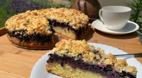 Ein saftiger Heidelbeerkuchen mit Streuseln, ein Stück auf einem Teller, daneben eine Tasse Tee.