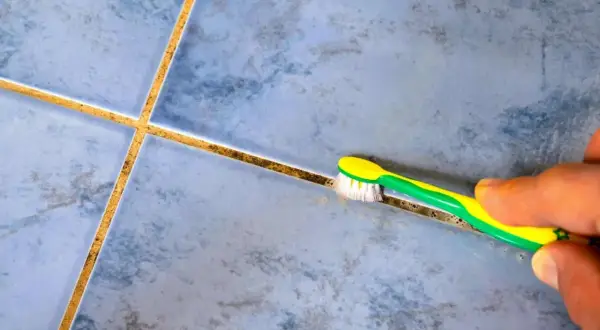 Une main utilise une brosse à dents pour blanchir le joint de carrelage sale, révélant la propreté cachée.