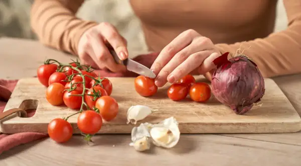 Hände schneiden Tomaten für selbstgemachten Ketchup. Zwiebel und Knoblauch liegen bereit.