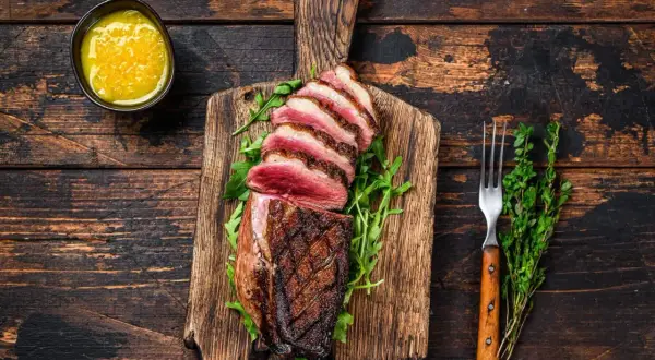 Perfekt gegarte Entenbrust mit rosa Kern, auf einem Holzbrett mit Rucola, dazu eine Schale mit Sauce und ein Gabel. Die Kerntemperatur der Entenbrust ist ideal.