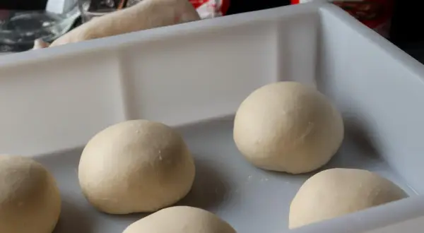 Mehrere Teigkugeln liegen in einer weißen Box, bereit für das Pizzateig einfrieren. Im Hintergrund sind Zutaten und eine Pizzatüte zu sehen.