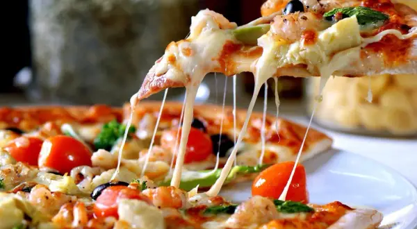 Rozciągający się ser z kawałka pizzy, na której widać krewetki, pomidorki i oliwki. Ciasto na pizzę wygląda apetycznie.