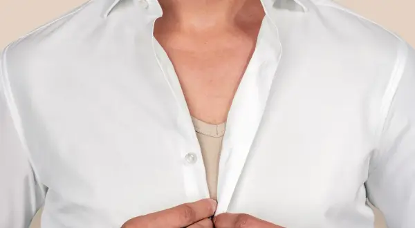 Mann schließt weiße Hemdknöpfe. Ein beiger T-Shirt ist unter dem Hemd sichtbar.