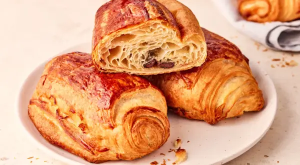 Złociste, chrupiące pain au chocolat na talerzu, jedno przekrojone, ukazujące puszyste wnętrze z kawałkami czekolady.