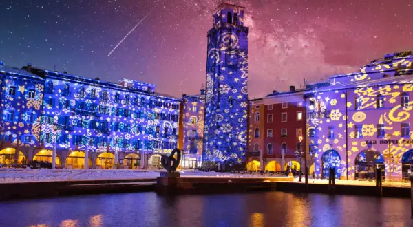Edifici illuminati da proiezioni natalizie sul lago di Garda a dicembre, con cielo stellato e una scia luminosa.