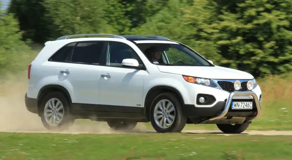 Białe Kia Sorento 1 zderza się z kurzem na drodze gruntowej, jadąc przez zielone pola.