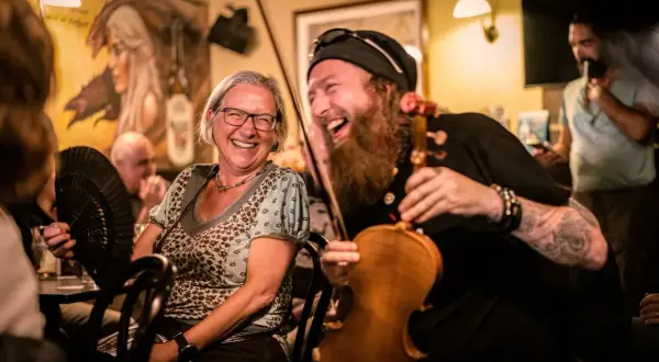Una mujer ríe mientras un violinista con barba toca. El ambiente es festivo, ¡qué buen craic!