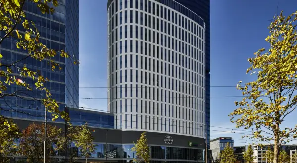 Nowoczesny budynek Crowne Plaza, część kompleksu Holiday Inn Warszawa, z charakterystyczną, falistą fasadą i zielenią wokół.