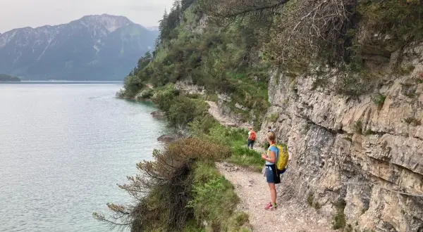 Wanderer genießen die Achensee Umrundung auf einem schmalen Pfad entlang steiler Felswände mit Blick auf den See und die Berge.