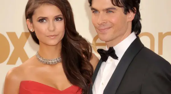 Nina Dobrev w czerwonej sukni i jej partner w smokingu, uśmiechnięci na ściance.