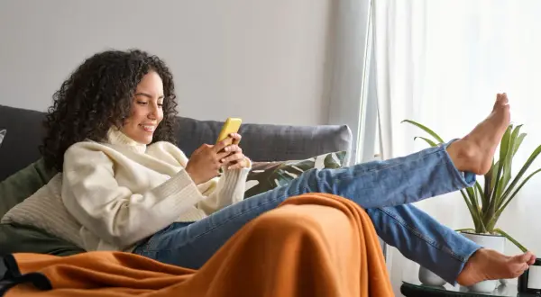 Frau mit lockigen Haaren entspannt sich richtig sitzend auf dem Sofa, schaut auf ihr Handy.