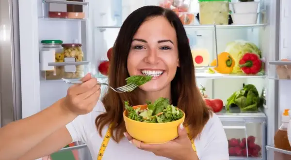 Uśmiechnięta kobieta je sałatkę z lodówki, wiedząc, że zdrowe jedzenie to klucz, jak przyspieszyć metabolizm.