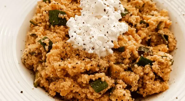 Un piatto di cous cous con zucchine e una generosa porzione di fiocchi di latte al centro, condito con pepe nero.
