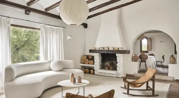 Salón acogedor con sofá curvo blanco, chimenea de piedra y mecedora de cuero. Un ejemplo de casas de campo sencillas y bonitas.