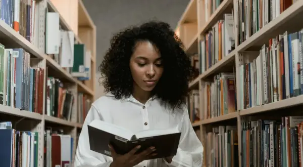 Młoda kobieta z burzą loków czyta książkę w bibliotece. Szuka inspiracji, co napisać we wniosku o stypendium szkolne.