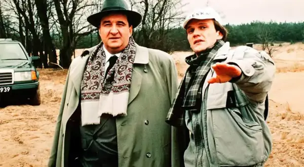 Dwóch mężczyzn w płaszczach i szalikach, jeden w kapeluszu, drugi w czapce. W tle drzewa i piasek. Nic śmiesznego.