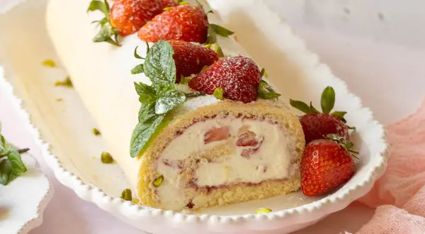 Eine köstliche Erdbeer Biskuitrolle, verziert mit frischen Erdbeeren, Minze und Pistazien, auf einem weißen Teller.