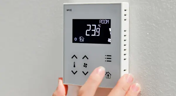 Dłoń naciska przyciski na termostacie, pokazując temperaturę 23.9°C. Dowiedz się, jak włączyć pompę ciepła, korzystając z intuicyjnego panelu sterowania.