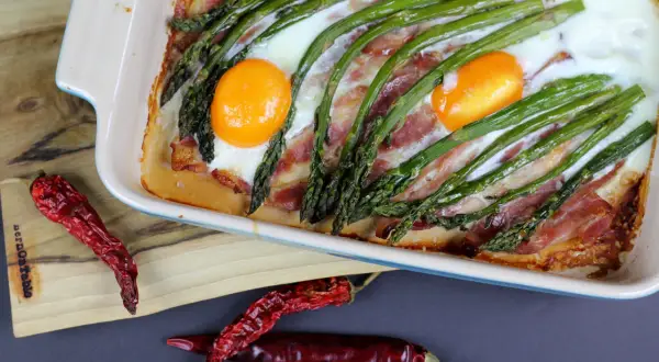 Pyszne śniadanie ze szparagami, jajkami i bekonem zapieczone w naczyniu. Obok leżą suszone papryczki chili.