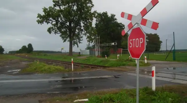Przejazd kolejowy z zaporami i znakiem STOP. Torowisko przecina mokrą drogę, a w tle widać drzewa i boisko.