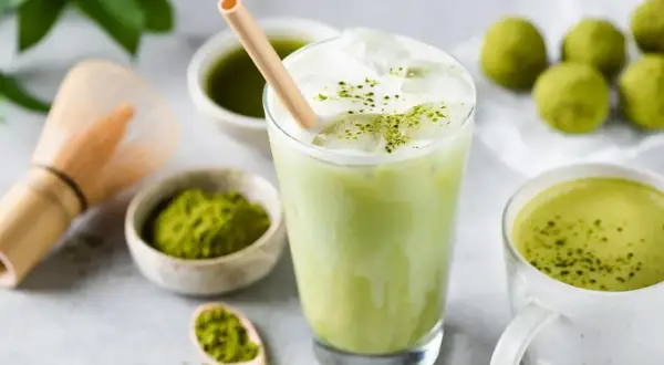 Orzeźwiający iced matcha latte z pianką i bambusową słomką, obok miseczki z matchą i tradycyjnej miotełki do jej przygotowania.