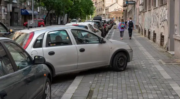 Samochody na chodniku utrudniają przejście. Dwie osoby idą wąskim chodnikiem, omijając przeszkody.