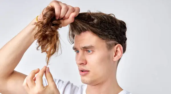 Mann mit blauen Augen betrachtet besorgt seine extrem kaputten Haare, die er in der Hand hält.