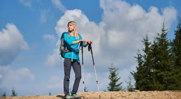 Kobieta z plecakiem i kijkami trekkingowymi na górskim szlaku. Zastanawia się, jak przymocować kijki do plecaka, by mieć wolne ręce.