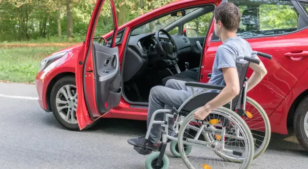 Uomo in carrozzina accanto a un'auto rossa con portiera aperta. Un disabile può avere due auto intestate per maggiore autonomia.
