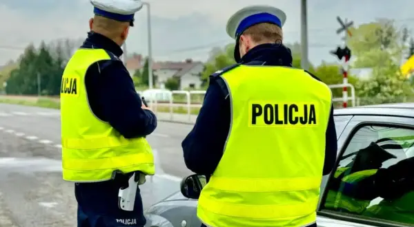 Dwóch policjantów w kamizelkach odblaskowych przy samochodzie. Jeden z nich trzyma broń. Prawdopodobnie zatrzymali kierowcę za przekroczenie prędkości o 50 km/h.
