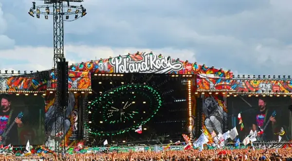 Ogromna scena festiwalowa z napisem "Poland Rock" i kolorowymi grafikami. Tłum ludzi czeka na koncert.
