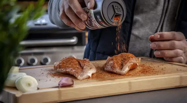 Ein Mann bestreut Hähnchenbrust mit einem magischen Gewürz-Rub auf einem Holzbrett. Zwiebeln und Knoblauch liegen daneben.