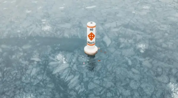 Bojka z napisem "NO BOATS" stoi na zamarzniętej tafli wody, ostrzegając przed ruchem łodzi.