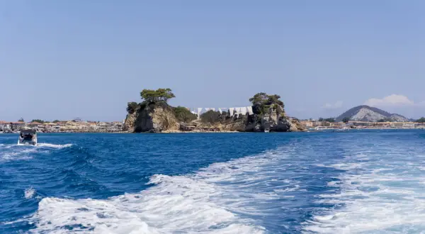 Idealna zakynthos pogoda sierpień: błękitne niebo, turkusowe morze i skaliste wysepki z drzewami. Widać łódź i plażę.