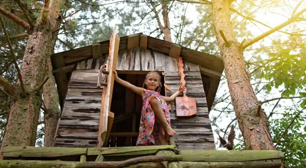 Dziewczynka w kolorowej sukience wita w swoim domku na drzewie. To inspiracja, jak zrobić domek na drzewie dla dzieci.