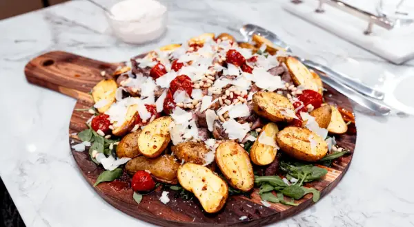 Biff Tagliata with roasted potatoes, tomatoes, pine nuts, parmesan, and arugula on a wooden board.