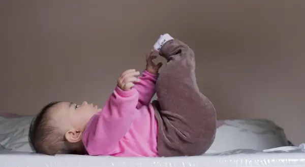 Niemowlę w różowym body i brązowych spodenkach leży na plecach, podnosząc nóżki. Pozycja ta może być pomocna w diagnostyce dysplazji stawu biodrowego.