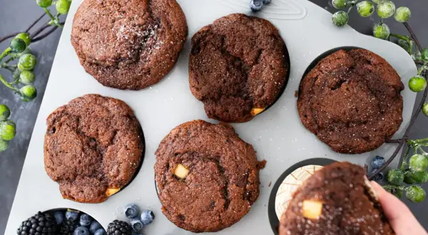 Fünf schoko muffins mit weißen Schokostückchen und Puderzucker in einer Muffinform, daneben Beeren und grüne Beeren.