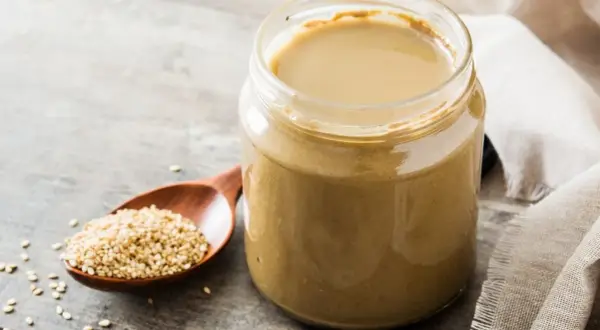 Słoik z gładkim, kremowym tahini obok drewnianej łyżki pełnej nasion sezamu. To właśnie tahini co to jest, czyli pasta z sezamu.