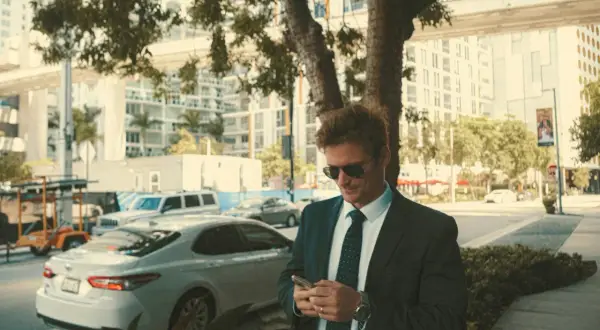 Mann im smart business dresscode, mit Sonnenbrille, blickt auf sein Handy. Im Hintergrund Stadtkulisse mit Autos.
