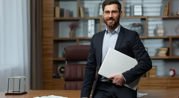 Uśmiechnięty dyrektor szkoły w garniturze, trzymający laptopa, stoi przy biurku w gabinecie z regałami pełnymi książek.