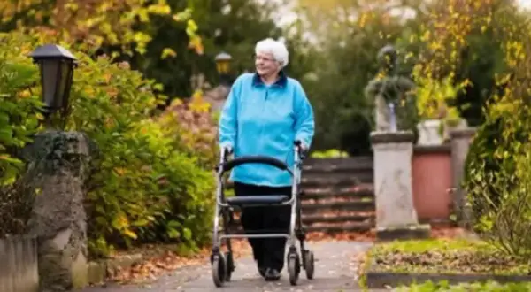 Elderly woman in blue jacket walks with a walker in a park with autumn foliage.